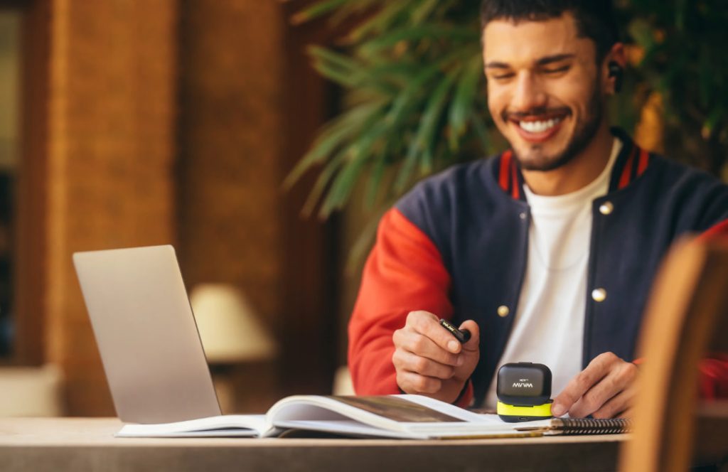 Homem sorrindo usa fone sem fio com notebook aberto e caderno.