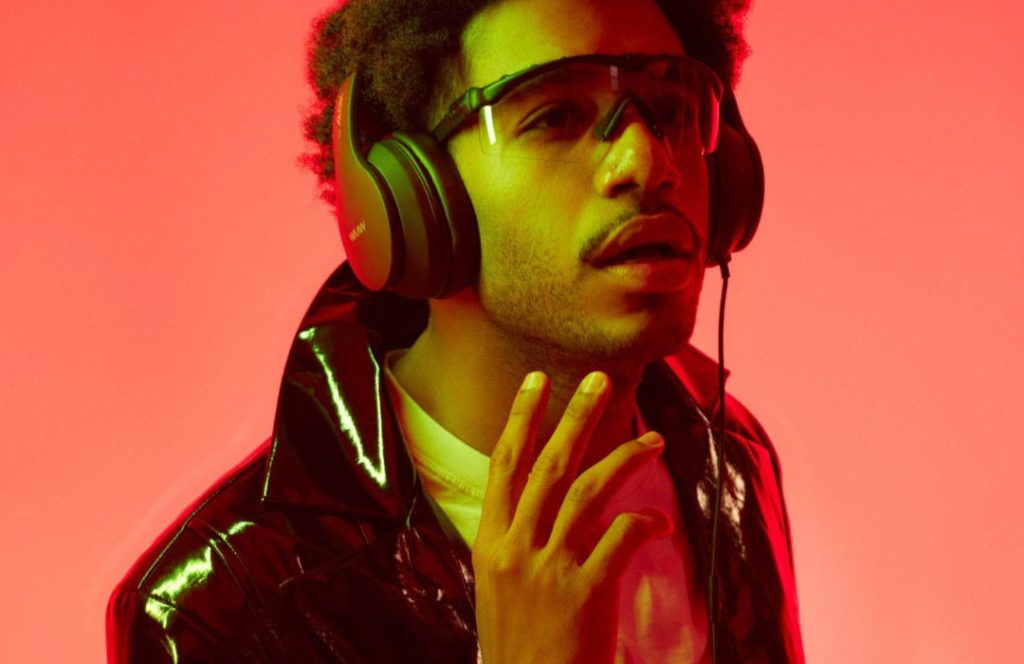 Homem com cabelo afro usa fone de ouvido preto da WAP e jaqueta brilhante, com fundo vermelho neon.